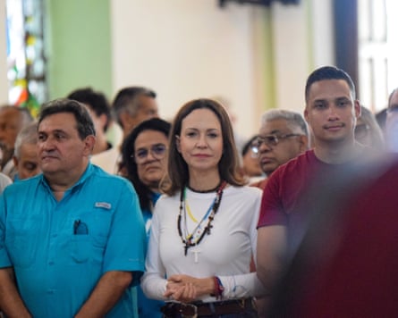 In a white, long-sleeved top and with a cross on a chain around her neck, María Corina Machado stands beside supporters with her hands clasped.