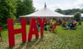 People sit in deckchairs on the grass outside a large white festival tent. In the foreground is the word HAY spelt out in large red letters erected on the grass