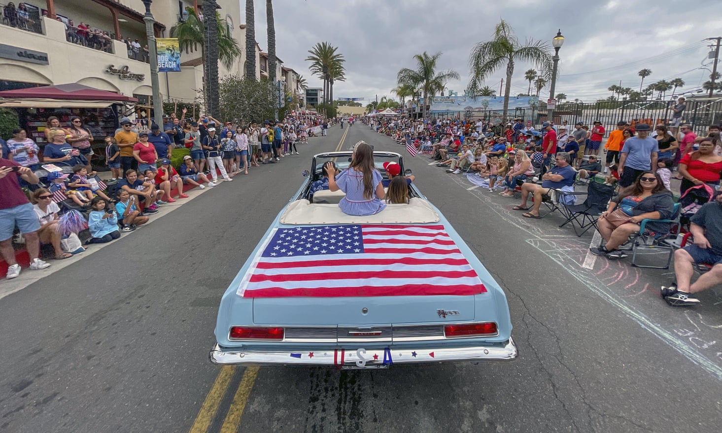 A long, older-model, powder-blue convertible with an American flag across the back half drives up a two-lane road crowded with people, with palm trees.