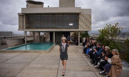 A model in black short-sleeved top and shorts with white criss-cross pattern, with pink shirt beneath and a straw hat, leads models past a pool on the roof of the modernist-style building