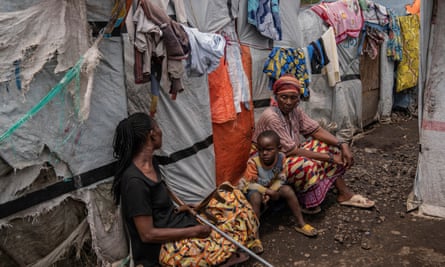 Two women and a child sit on the ground in front of huts