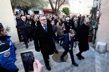 Nicolas-Jacques Charrier, who is in his mid-60s and dressed in a dark coat, walks across a stone square holding the hand of a young boy who holds the hand of a blond-haired woman to his other side. A large group of people are following them.