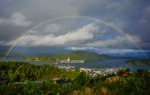 A rainbow spans over a large military ship in a fjord, an islet behind it