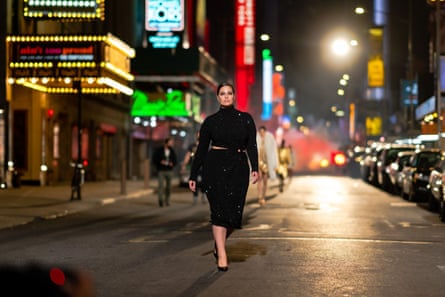 Plus size model Ashley Graham dressed in black walking along a New York street lit up with colourful neon