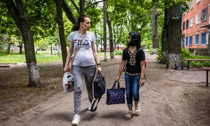 A young woman with the first signs of a baby bump talks to a young girl who is walking with her, carrying bags