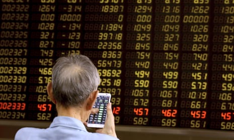 A man looks at a phone in front of a screen showing stock prices