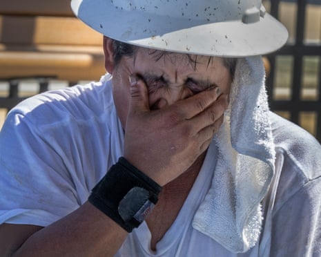 A man in a hard hat wipes sweat from his eyes with his hand, a towel around his neck