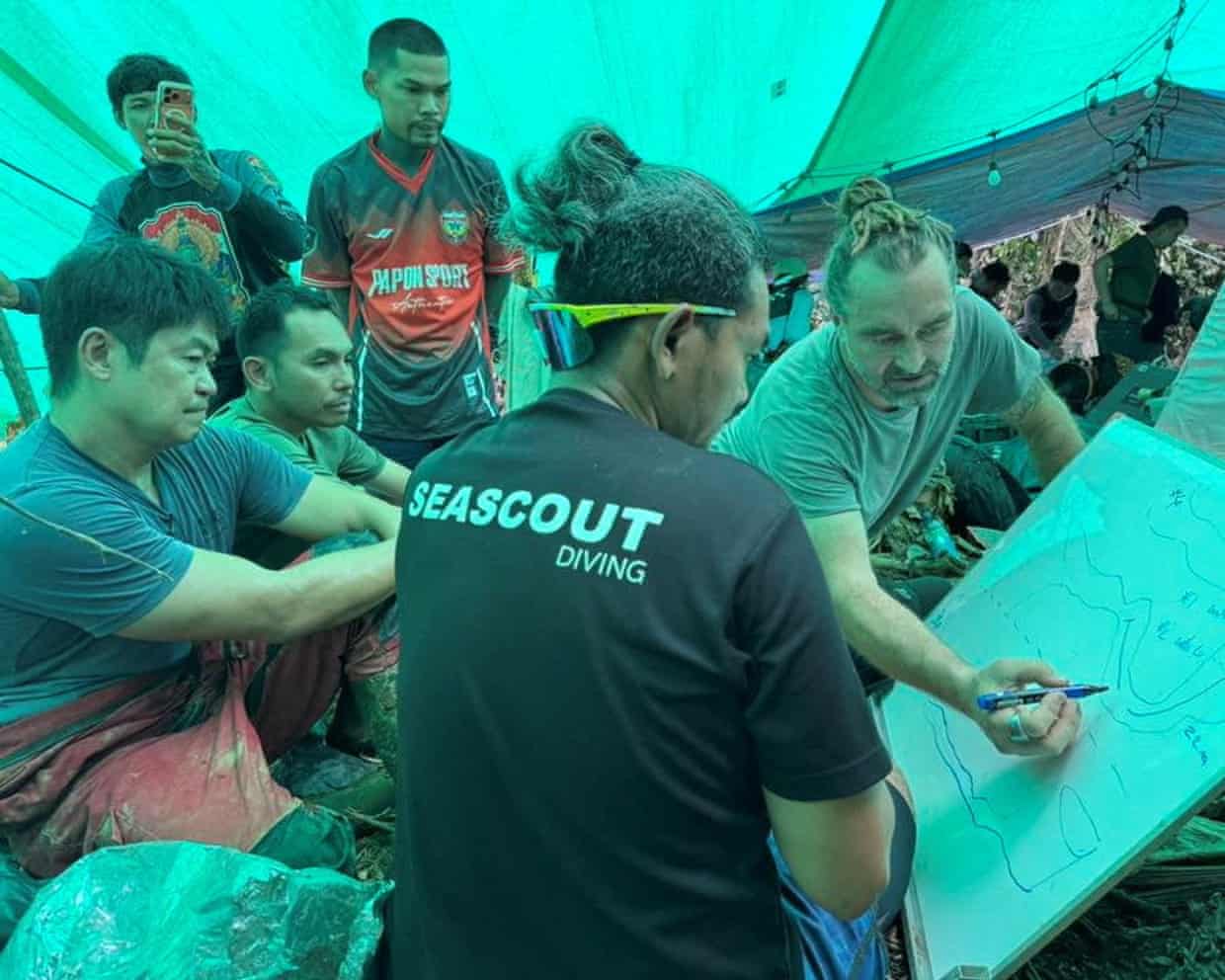 People gather around a man drawing on a whiteboard under blue tarpaulin