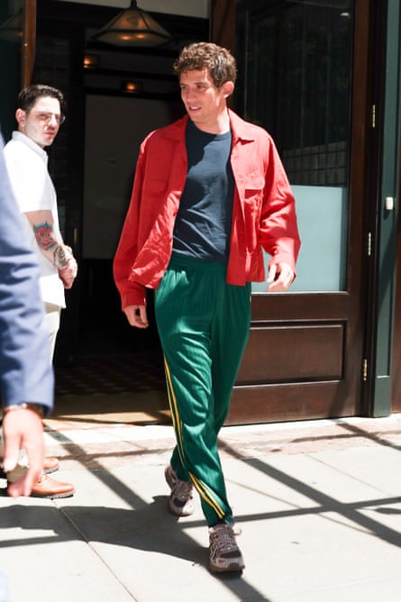 Josh O’Connor in green tracksuit bottoms with yellow stripes, trainers, and a black T-shirt with a red open shirt over it