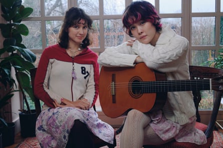 Mei Semones leans on an acoustic guitar while sat next to Liana Flores in front of a window