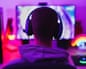 Rear view of a young man wearing a headset, gaming at a computer in a neon-lit room