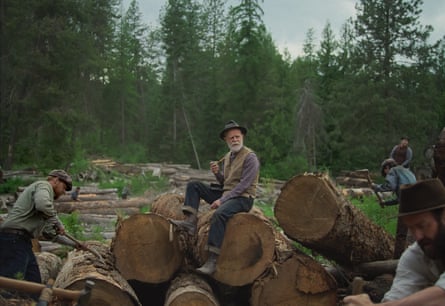 William H Macy with white beard, pipe and hat, sitting on logs while men work around him, in Train Dreams