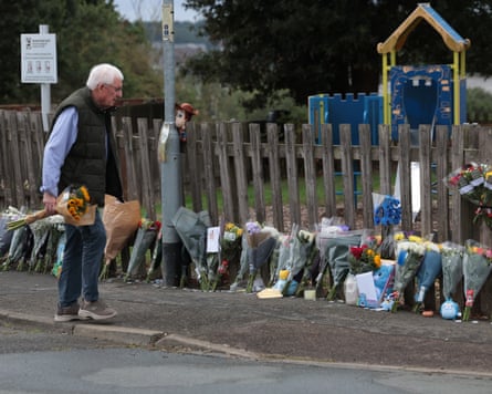 A man with white hair holding a bunch of flowers stands on the path beside the playground and looks at the flowers lining the length of the fence