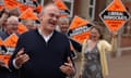 Ed Davey campaigns in Oxfordshire: he stands in front of a group of people waving bright orange placards that read Liberal Democrats: winning here. He wears a dark jumper over a white shirt and looks animated, with his hands raised.