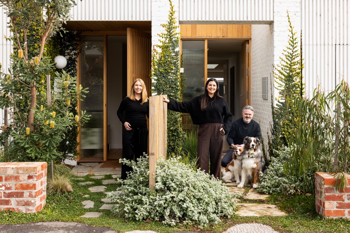 Three people and two dogs are seen in the yards of two townhouses, talking over the fence