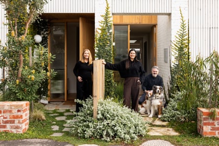 Three people and two dogs are seen in the yards of two townhouses, talking over the fence