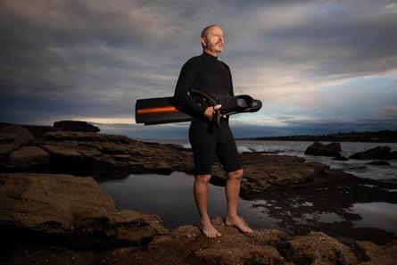 Marine biologist and shark expert Prof Rob Harcourt stands in a wetsuit holding flippers at dusk on the rocks at Bondi Beach