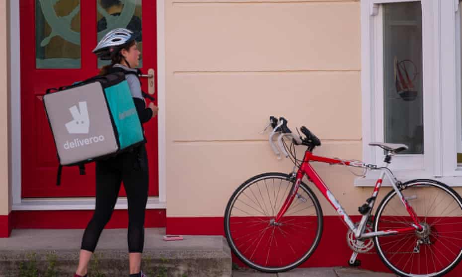 young woman delivering Deliveroo meal to a house with her bicycle alongside