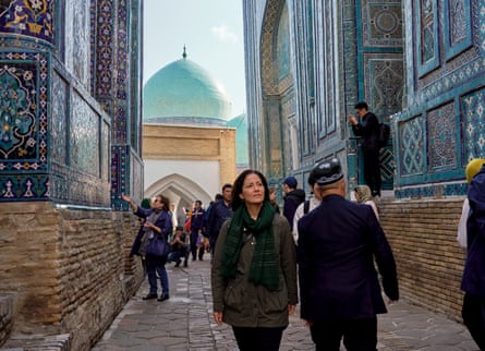Mishal Husain wearing a dark green scarf and jacket, walking between two intricately tiled buildings, with blue domed buildings seen at the end of the alleyway