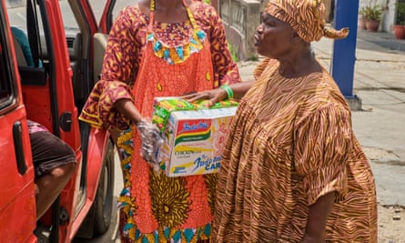 An African woman in matching dress and headscarf carries a large box of instant noodles