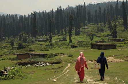 Two women in shalwar kameez walk along a path in a green rural setting towards some trees