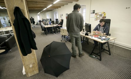 A photo taken in a large interior room with a low ceiling and neat wooden rafters, florescent lights, gray industrial carpeting, and large wooden tables. A black raincoat hangs on a wooden post, and a black umbrella is open in the foreground. Two white men appear to chat, one seated at one of the desks and the other standing alongside it.