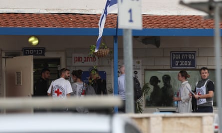 Representatives of the ICRC arrive at the Ofer military prison in the West Bank: three men and a woman, all wearing white tabards with red cross symbols on them, are seen about to walk through a door with several others. They are entering a building with a breezeblock front and tiled roof over the porch; there are signs in Hebrew and a blue and white Israeli flag billows.