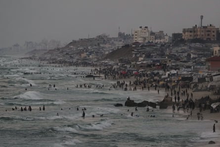 People stand and swim in the sea and walk on a beach that is full of tents reaching almost to the water’s edge. The city is visible beyond the beach, under a grey sky.