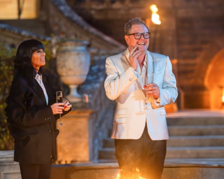 An image of Claudia Winkleman and comedian Alan Carr on the set of the BBC show The Traitors. They are standing outdoors at night in front of a stone staircase at Ardross Castle. Claudia is on the left, wearing a black blazer and holding a glass of champagne. Alan Carr is on the right, wearing a light-coloured suit jacket and glasses, laughing while also holding a glass of champagne.