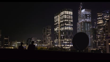 Two silhouetted people stand on a rooftop overlooking a city skyline lit up at night