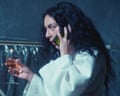 A dark haired woman in a dressing gown holds a drink in her hand while she talks on a phone with the other.