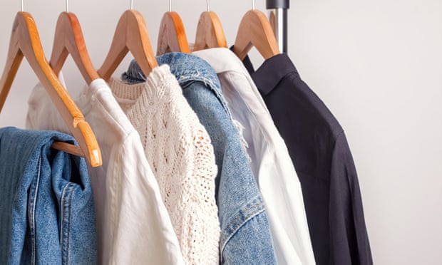 A selection of clothes including some jeans, white tops, a denim jack and a dark shirt, hanging on wooden coat hangers on a rack