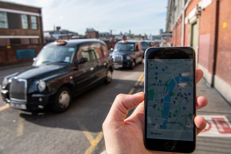 A phone displaying the Uber app in front of a taxi rank at Waterloo station in London.