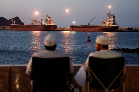 Two men in the foreground sit facing the two tankers, which are anchored across the other side of the port