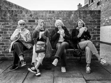 Four women laughing and posing for the camera as they enjoy chips on a bench – image is black and white