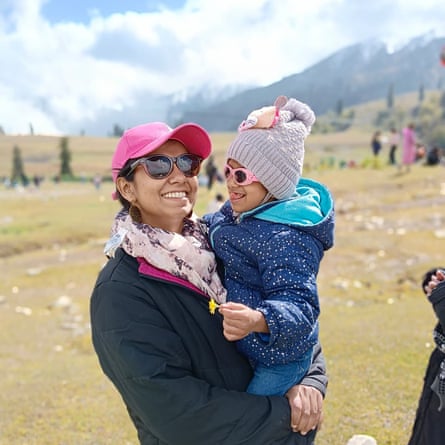 A smiling woman standing outside holding a child who carries a flower. Both have a hat and sunglasses on