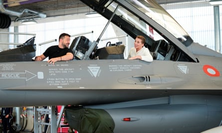 Volodymyr Zelenskiy blickt zurück auf Mette Frederiksen, die in einem F-16-Kampfflugzeug in einem Flugzeughangar sitzt