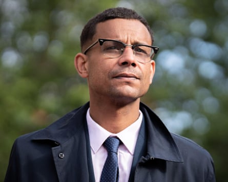Daniel Kebede wearing glasses, a dark coat and a navy tie standing against a backdrop of trees.