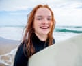 A girl with red hair walks along a beach with surfboard under her arm.