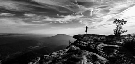 A black and white photo showing a man on the edge of a rock looking into the wilderness.