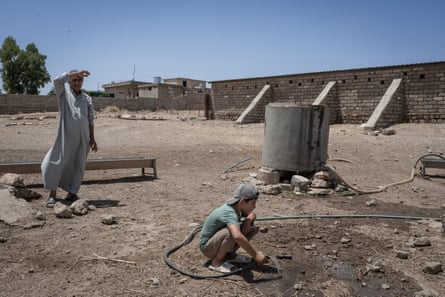 A man stands next to a small boy crouching holding a hose in a brick-walled compound