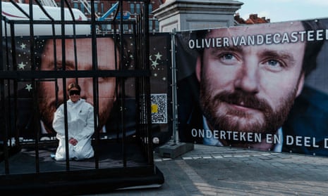 A woman blindfolded sits in a cage next to a large poster with man's face on it in protest at his imprisonment.