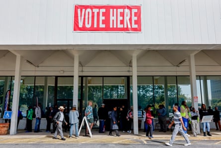 People wait in a building’s shade beneath a large “vote here” sign.