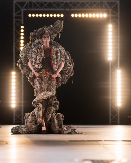 A woman in an outfit with oversized hat and sleeves, and ruffles from the knees down, in front of a square structure