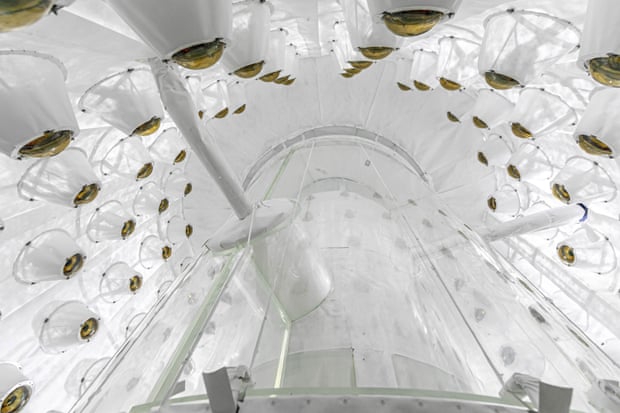 An all-white chamber, seen from a low angle, is lined with dome-shaped devices, all aimed at a clear cylinder.