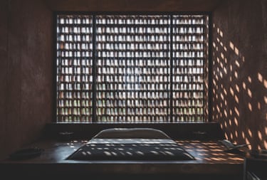 A bed sits before a large latticed window casting geometric shadows across a dim room