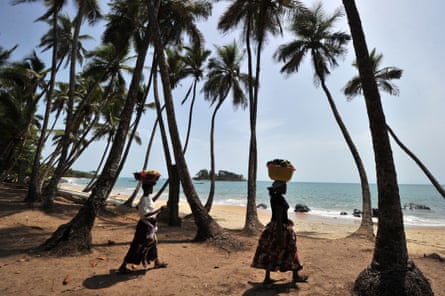 two women carry baskets on their heads by a palm tree-fringed beach