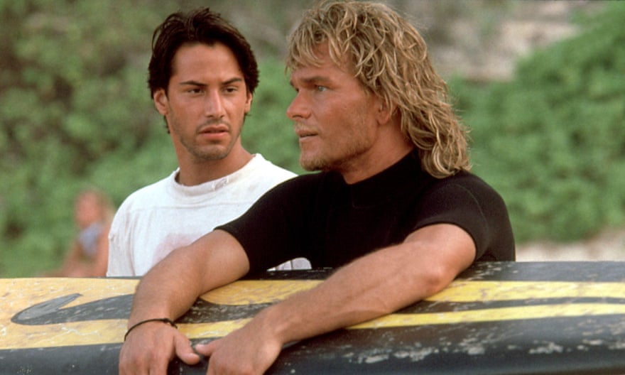 Keanu Reeve in a T-shirt holding a surfboard watches Patrick Swayze in a rash vest