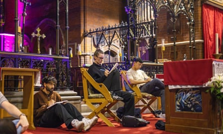 three people sit writing – one on the floor and two in wooden chairs – in front of a church altar