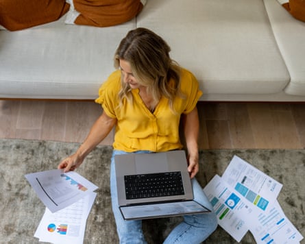 A person with long hair and wearing a short sleeved yellow shirt and pale blue jeans sits on the floor with her back against the sofa while holding a laptop and looking at marketing material